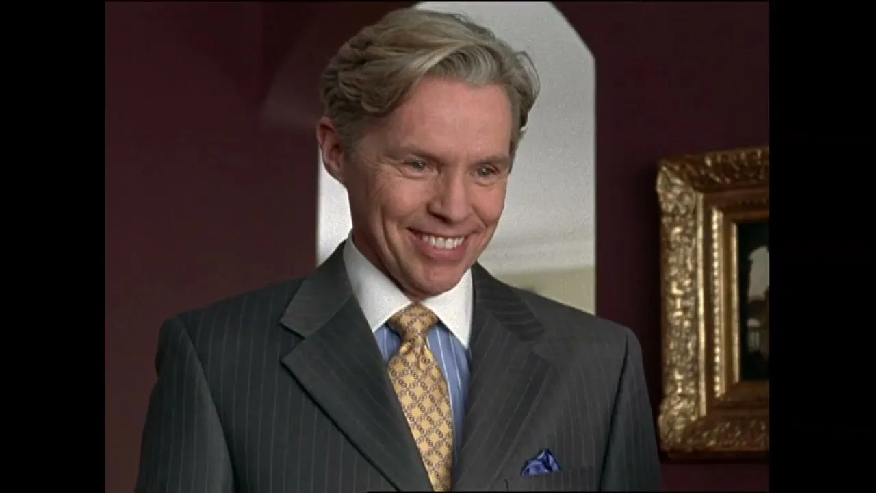 A smiling man in a pinstripe suit and patterned tie looks down and to the right in an interior setting.