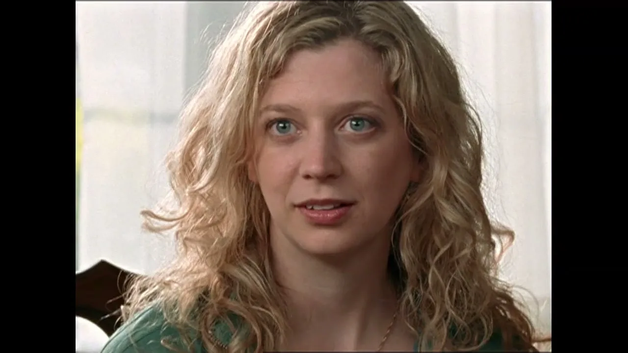 A young blonde woman with curly hair and blue eyes looks slightly off-camera, listening intently in an interior setting.