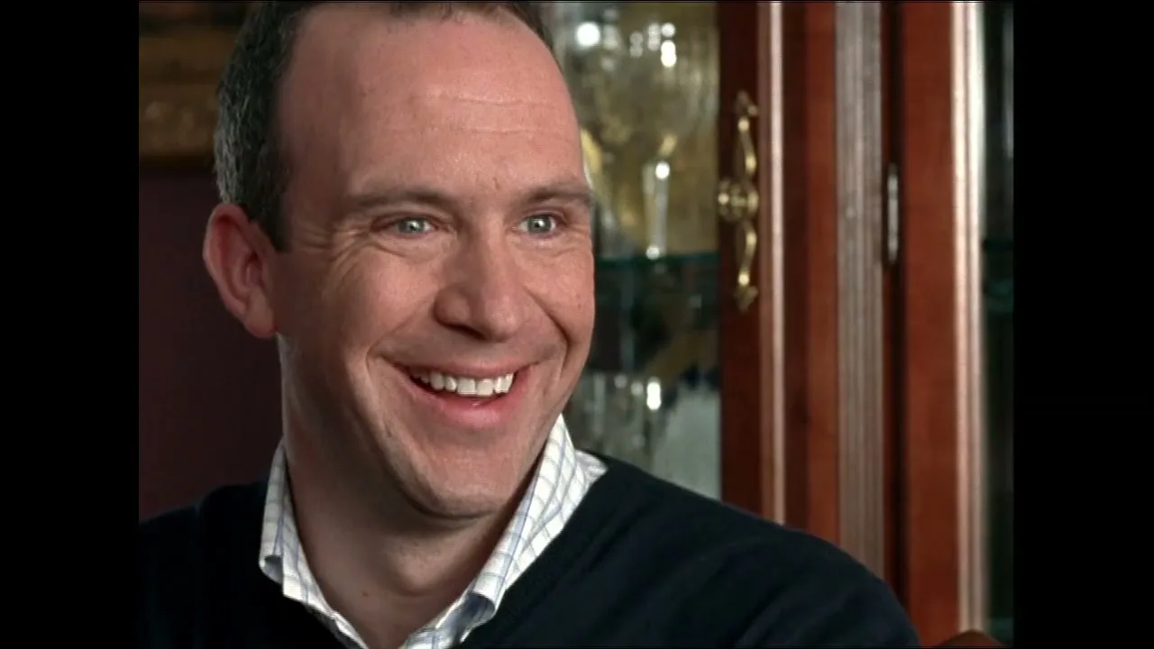A man with short hair and a collared shirt smiles, revealing white teeth, in an interior setting with a wooden cabinet behind him.