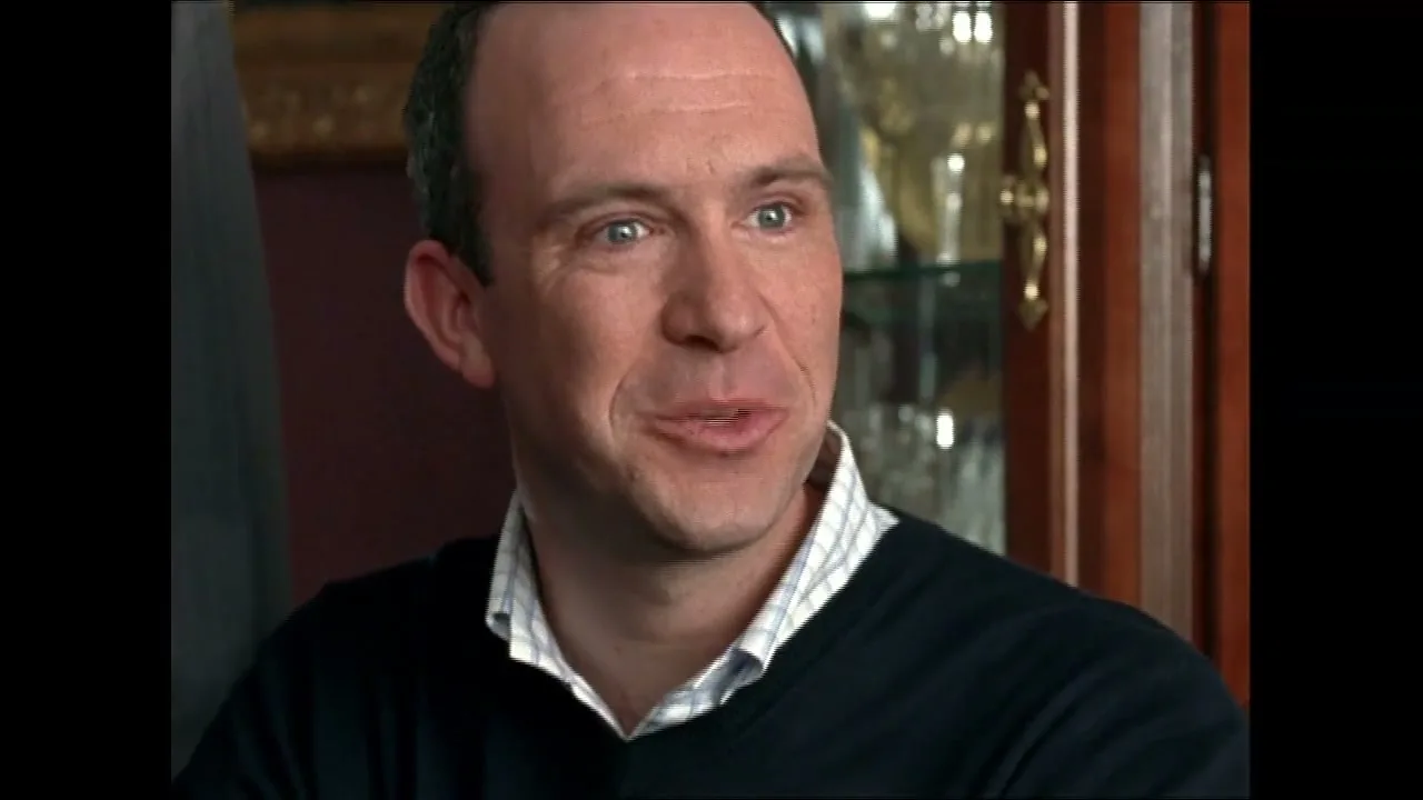 A balding man in his 30s, wearing a dark sweater over a plaid shirt, smiles and speaks in an interior setting.