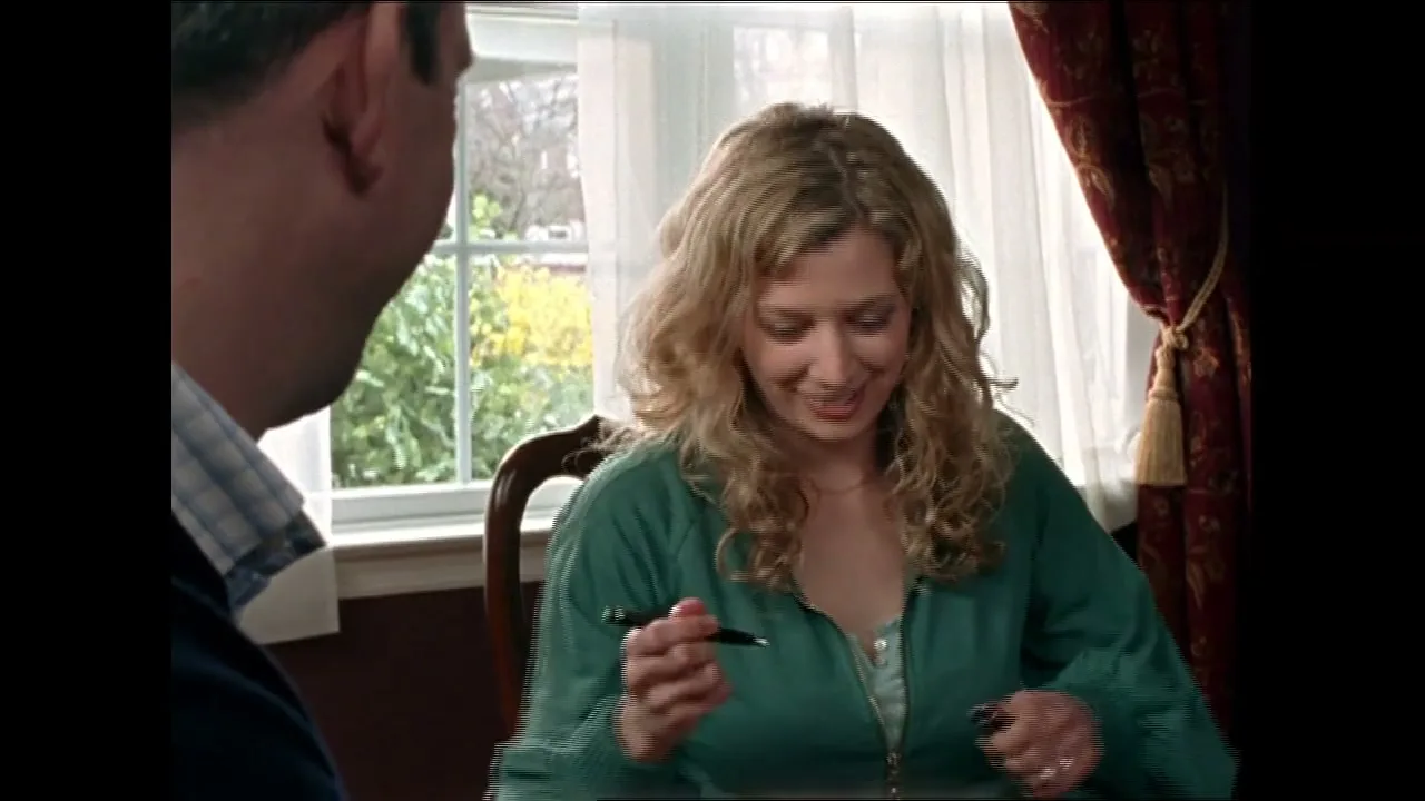 A blonde woman in a green top smiles and signs a document while sitting across from a man in a well-lit room.