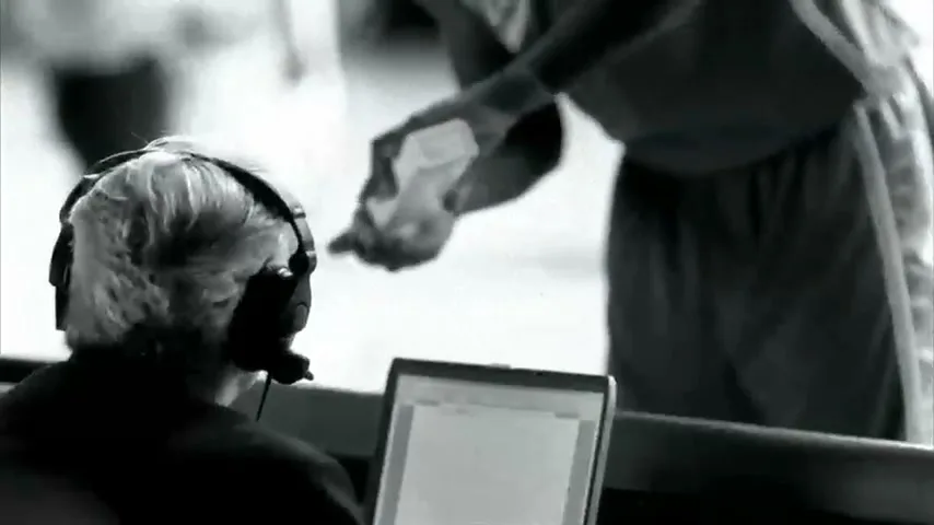 A person with headphones and a laptop watches a basketball player in the background, who is preparing to throw chalk into the air.