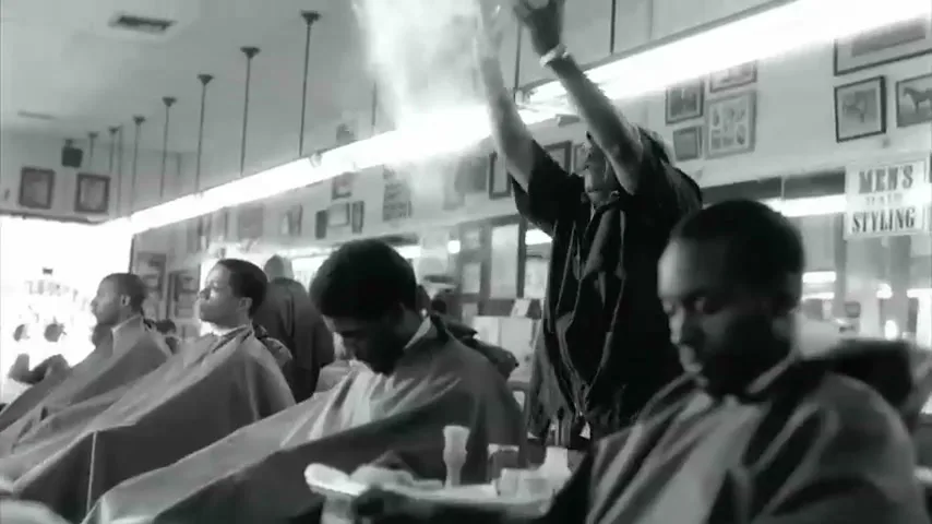 A barber in a flat cap and dark shirt throws talcum powder into the air in a busy barbershop, with several men in barber capes seated.