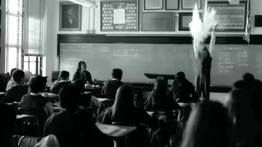 A young male student in a classroom throws a piece of paper into the air, creating a chalk dust cloud, while other students and a teacher are present.