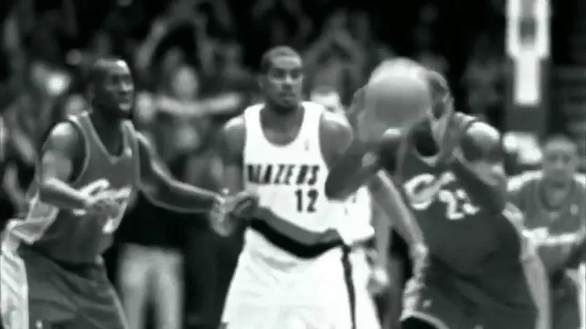 A black and white shot of basketball players on a court, with one player in a white jersey with "BLAZERS 12" on it, and another player in a dark jersey dribbling a basketball.