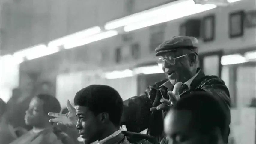 An older Black man in glasses and a cap gestures animatedly while cutting a young Black man's hair in a busy barbershop.