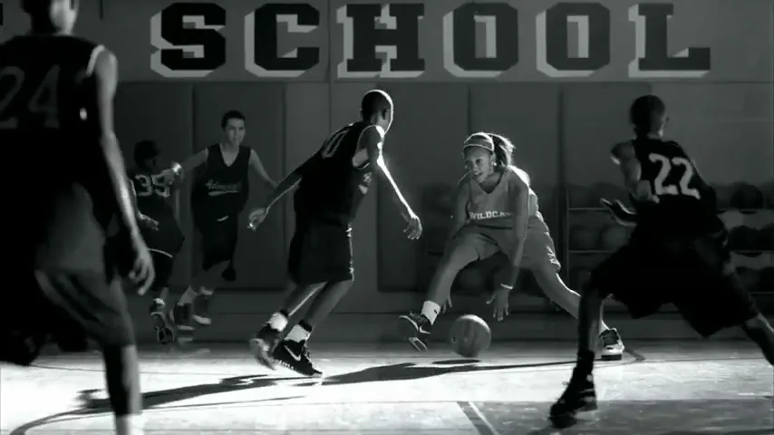 A female basketball player in a jersey dribbles the ball on a court, surrounded by male players, in a school gym.