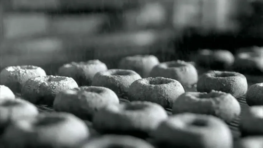 A close-up shot shows many small, round, chalk-dusted donuts on a conveyor belt, moving from left to right in a factory setting.