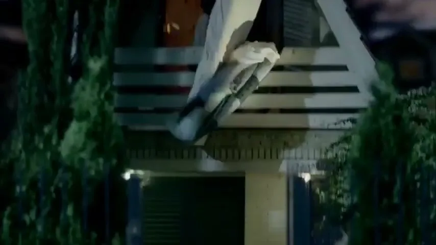 A person in a white sheet and blue backpack climbs out of a window on the second story of a house at night.
