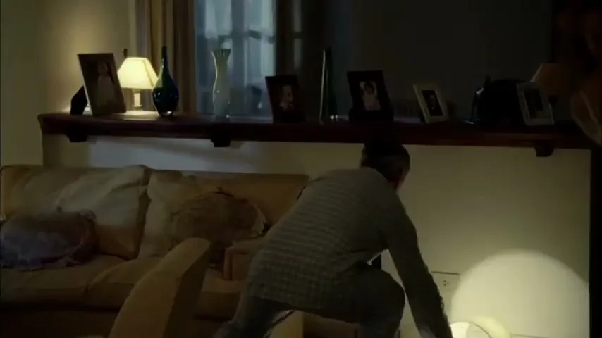 A man in pajamas frantically searches for something on the floor, while a woman in a robe enters the frame in a dimly lit living room.