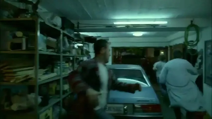 Three men in casual clothes run through a cluttered garage towards a car, one man in a plaid shirt reaches for a tool board.