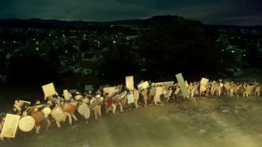 A large group of people, dressed in ancient warrior attire, march in a long line across a dark, barren hillside overlooking a city at night.