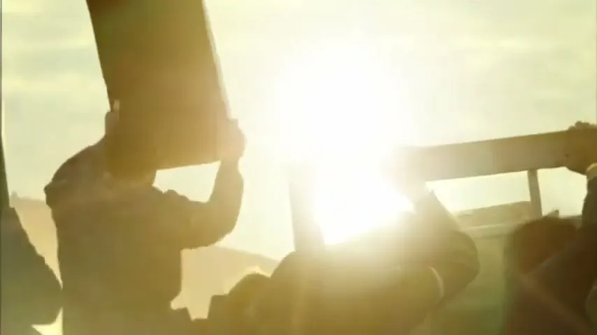 Silhouetted people in a truck bed hold up signs, facing a bright sun in a wide shot.