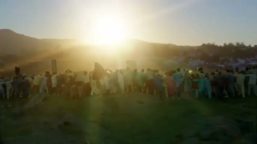 A large crowd of people, dressed in various light-colored clothing, stand together on a grassy hill at sunrise, with mountains in the background.