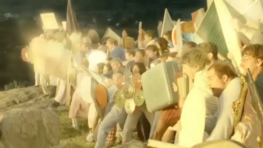 A large group of diverse people, dressed in casual clothes, are seen from the side, holding various household items as shields, as they push forward on a grassy hill at dawn.