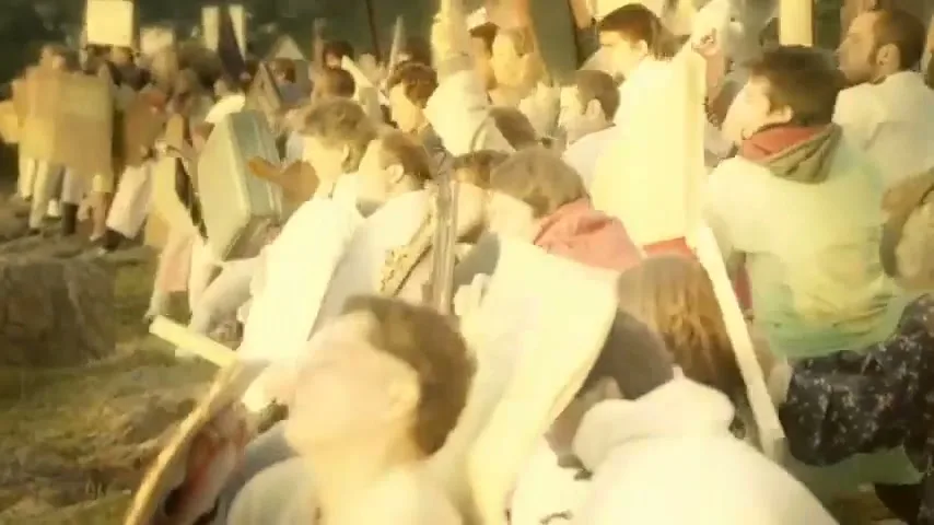 A large group of diverse people, dressed in white and holding signs, gather outdoors in a protest or demonstration during golden hour.