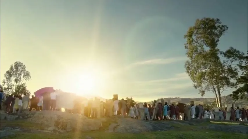 A diverse crowd of people, dressed in various outfits, stands on a rocky hill, facing a bright sunrise in a wide shot.