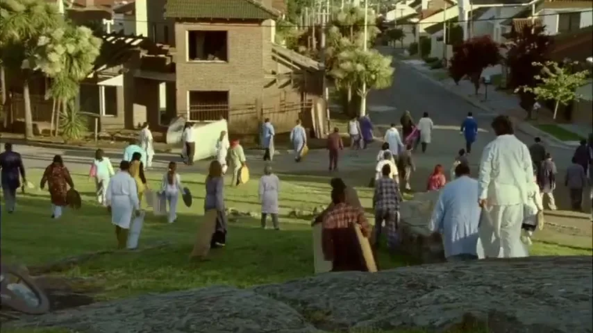 A diverse group of people, dressed in various work uniforms, walk across a grassy hill in a suburban neighborhood, carrying their belongings.