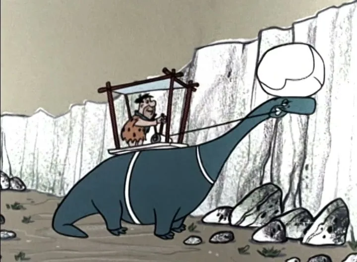 Fred Flintstone, a cartoon caveman, rides on the back of a large blue dinosaur, which is carrying a rock on its head, in a prehistoric, rocky landscape.