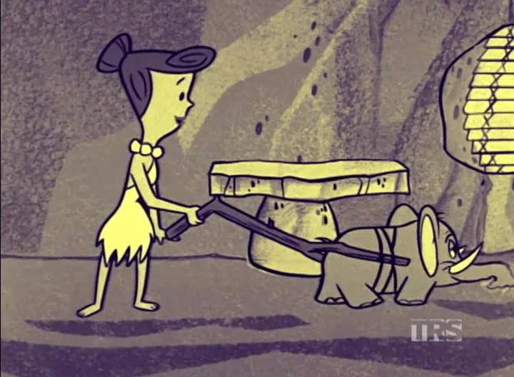 Wilma Flintstone, a cartoon woman with a bone in her hair and a cavewoman dress, uses a stick to operate a small elephant vacuum cleaner in a cave.