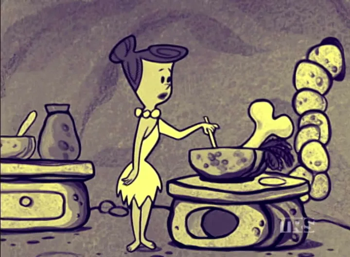 Betty Rubble, a cartoon character, stands in a cave kitchen, stirring a large bowl with a bone in it.