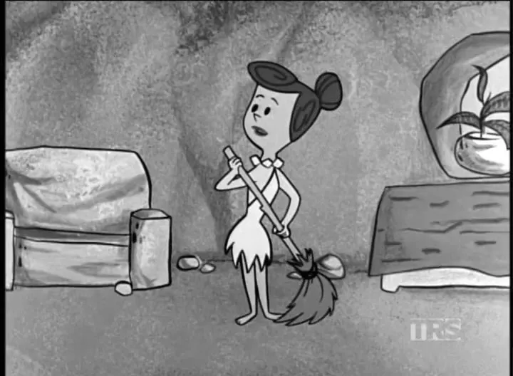 Betty Rubble, a cartoon character, stands in a cave-like living room, holding a broom and looking up with a concerned expression.