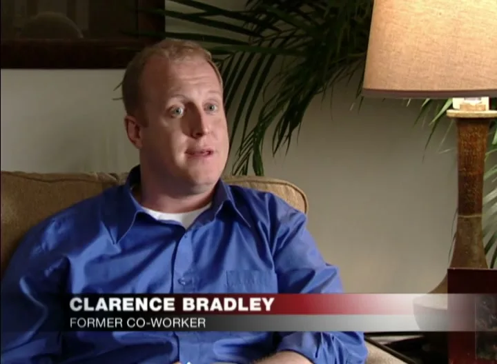 A man in a blue shirt sits on a couch, speaking to the camera, with a plant and lamp in the background.