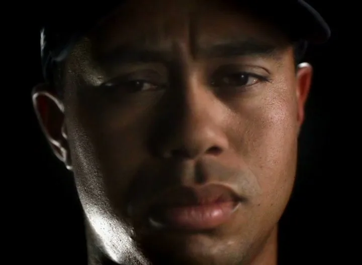 Tiger Woods, a male golfer in his 30s, wears a dark cap and shirt, looking intently at the camera in a dark studio.