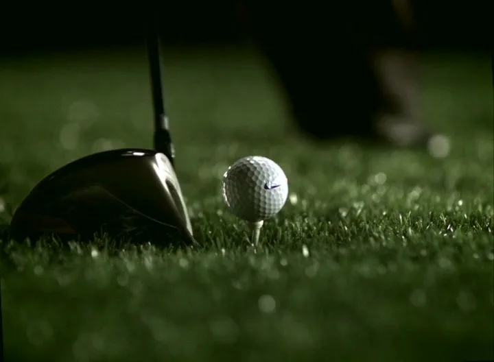 A golf club is positioned next to a golf ball on a tee, ready to be hit on a dark green golf course.