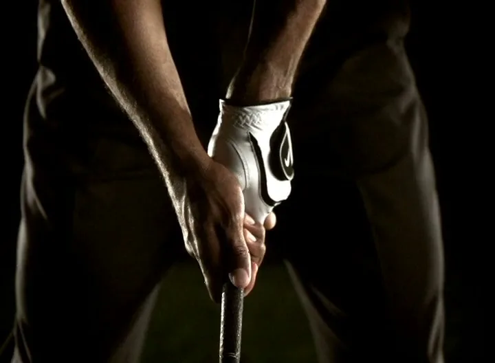 A golfer's hands, one gloved, grip a golf club in a dark, dramatic setting, preparing for a swing.