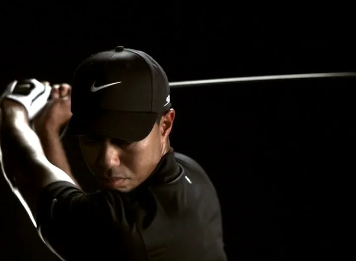 A male golfer in a black cap and shirt, with a white glove, holds a golf club, preparing to swing in a dark studio.
