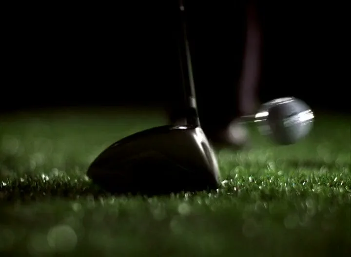 A golf club swings and hits a golf ball off a tee on a patch of green grass against a dark background.