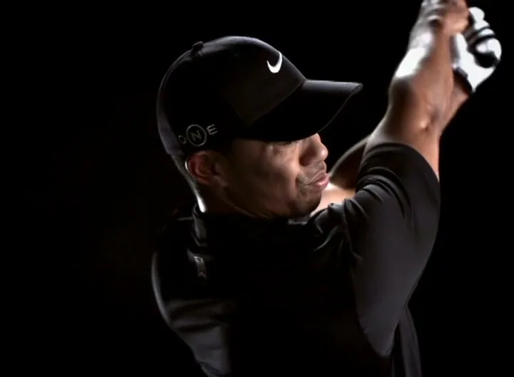 Tiger Woods, a male golfer in his 30s, wearing a black cap and shirt, swings a golf club in a studio void.