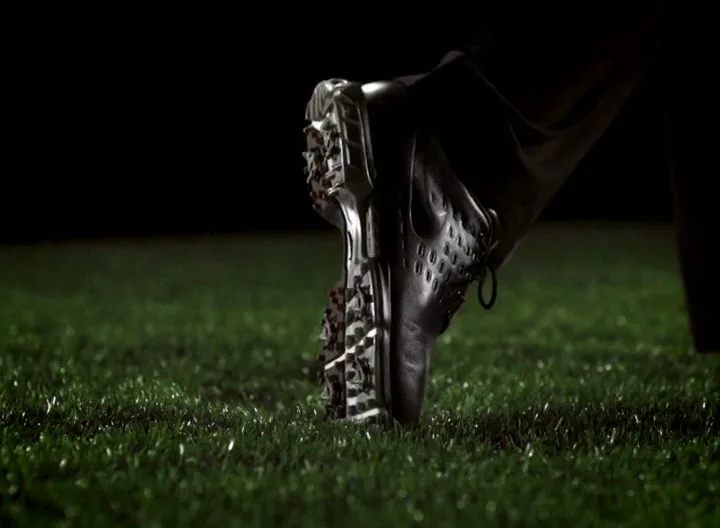 A golf shoe with spikes is seen in a close-up shot, pressing into green grass against a dark background.