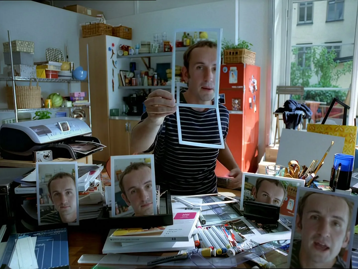 A man in a striped shirt sits at a messy desk, holding up a white frame and then a photo of himself, surrounded by other photos of his face.