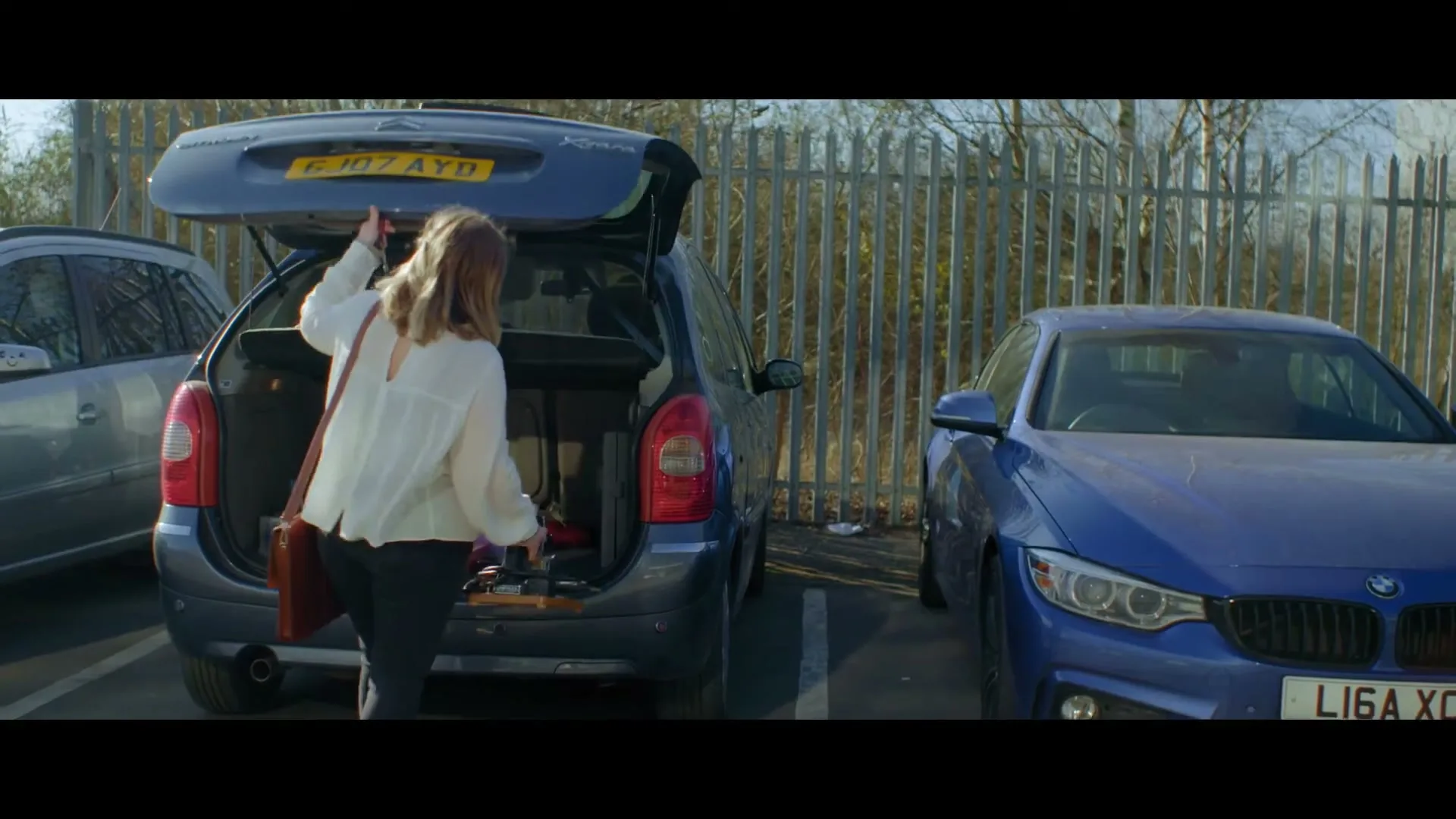 A woman with brown hair, wearing a white shirt and black pants, puts items into the open trunk of a blue car in a parking lot.