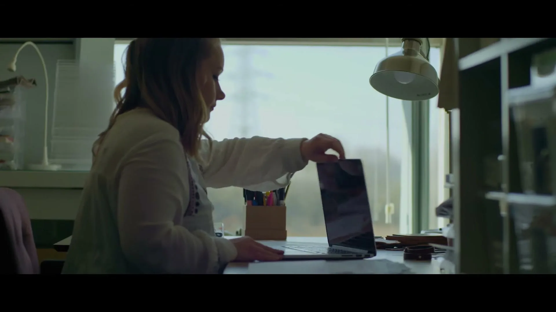 A woman with blonde hair, wearing a white top, sits at a desk by a window, opening a laptop in an office.