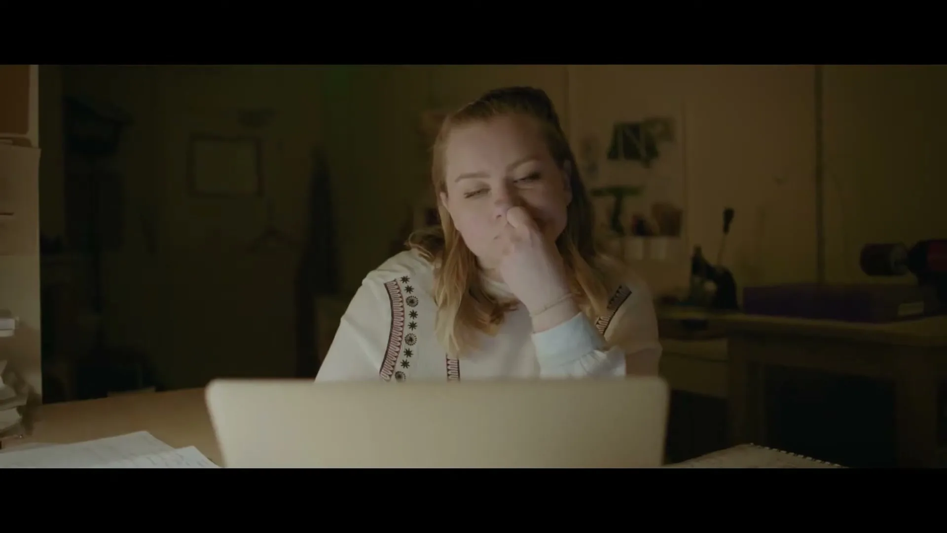 A young woman with blonde hair, wearing a white embroidered shirt, sits at a desk, looking tired and rubbing her nose while working on a laptop in a dimly lit room.