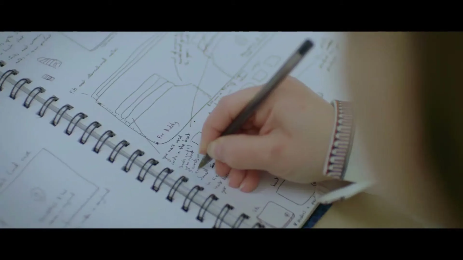 A person's hand writes notes and sketches in a spiral-bound notebook, seen from over their shoulder.