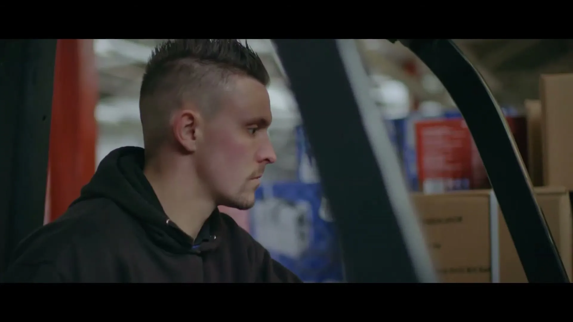 A young man with a mohawk and black hoodie sits in a forklift, looking intently to the right in a warehouse.