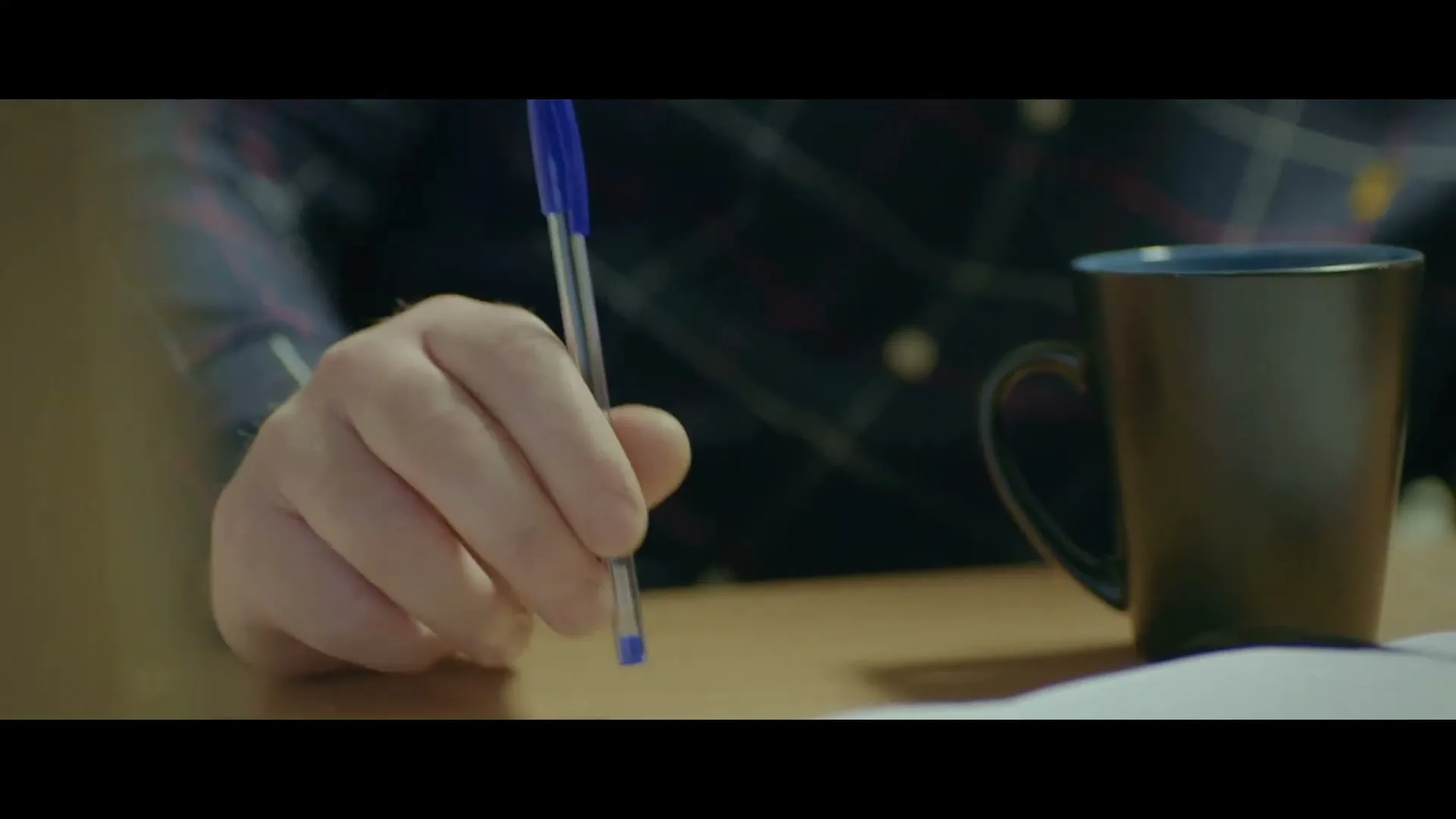 A man's hand holds a blue pen, tapping it on a table next to a steaming mug and a piece of paper.