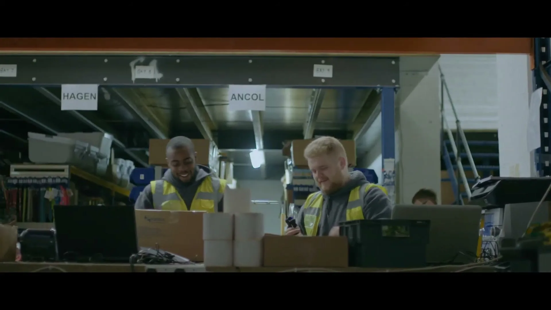 Two male warehouse workers in high-vis vests smile and laugh while working at a packing station in a busy warehouse.
