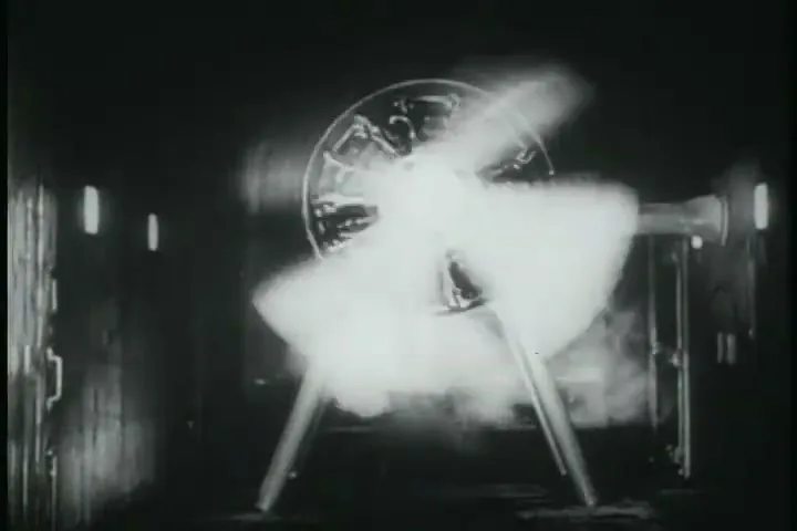 A black and white shot shows a large, glowing propeller-like object spinning rapidly in a dark, industrial setting.