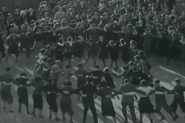 A large group of people, dressed in dark clothing, hold hands and dance in a circle on a paved outdoor area, viewed from above.