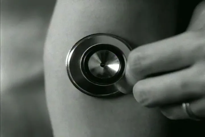 A close-up shot shows a person's hand holding a stethoscope to another person's bare arm, in a black and white, high-contrast setting.