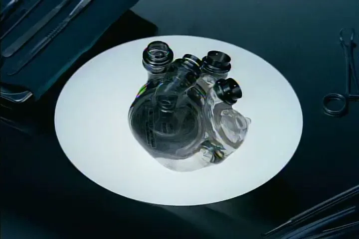 A futuristic, transparent artificial heart sits on a glowing white disc, surrounded by surgical tools on a dark surface.