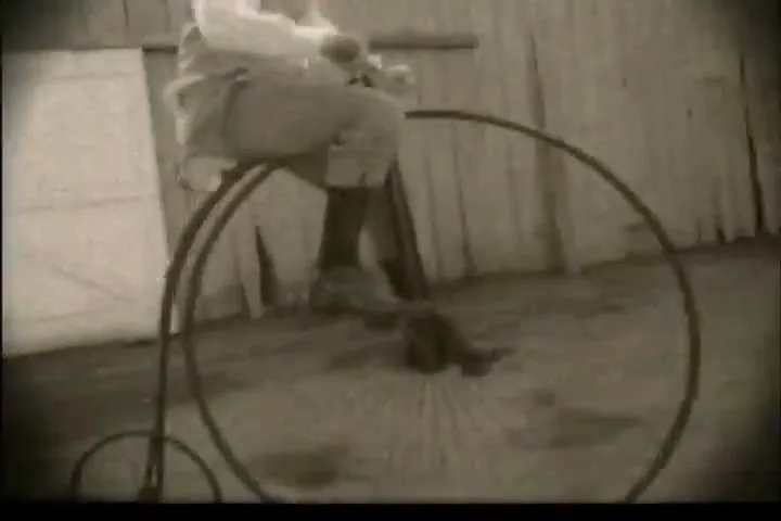 A person in old-fashioned clothing rides a penny-farthing bicycle on a dirt path next to a wooden building, filmed in sepia tone.