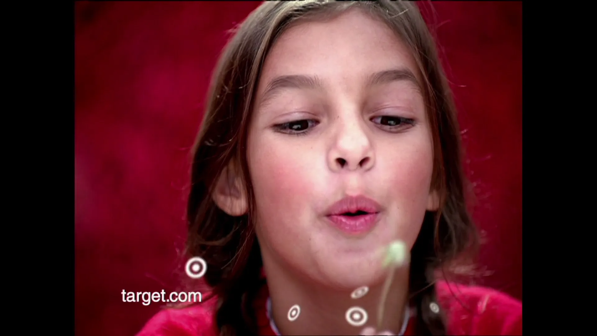 A young girl with brown hair in pigtails blows on a dandelion, creating floating Target bullseye logos against a red background.