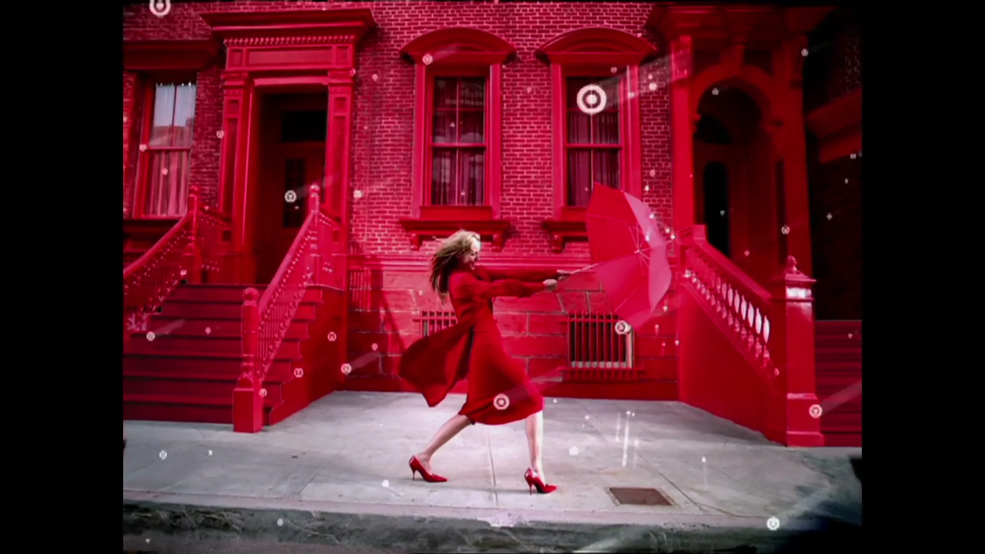 A woman in a red coat and heels struggles with a red umbrella against a strong wind, with a red brick building in the background, as white bullseye shapes fall like rain.