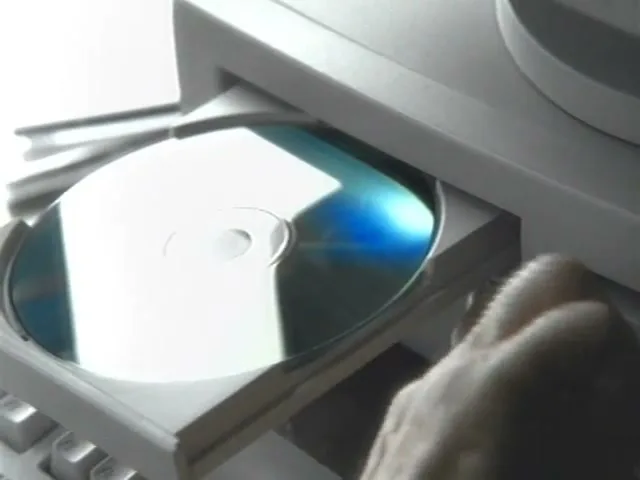 A hand inserts a CD into a desktop computer's CD-ROM drive, which then closes, on a desk.